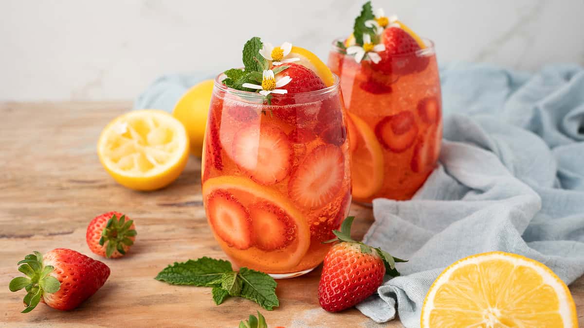 Two glasses of strawberry and lemon infused water with ice, garnished with mint and flowers, sit on a wooden surface with fresh strawberries, lemon halves, and mint leaves nearby.