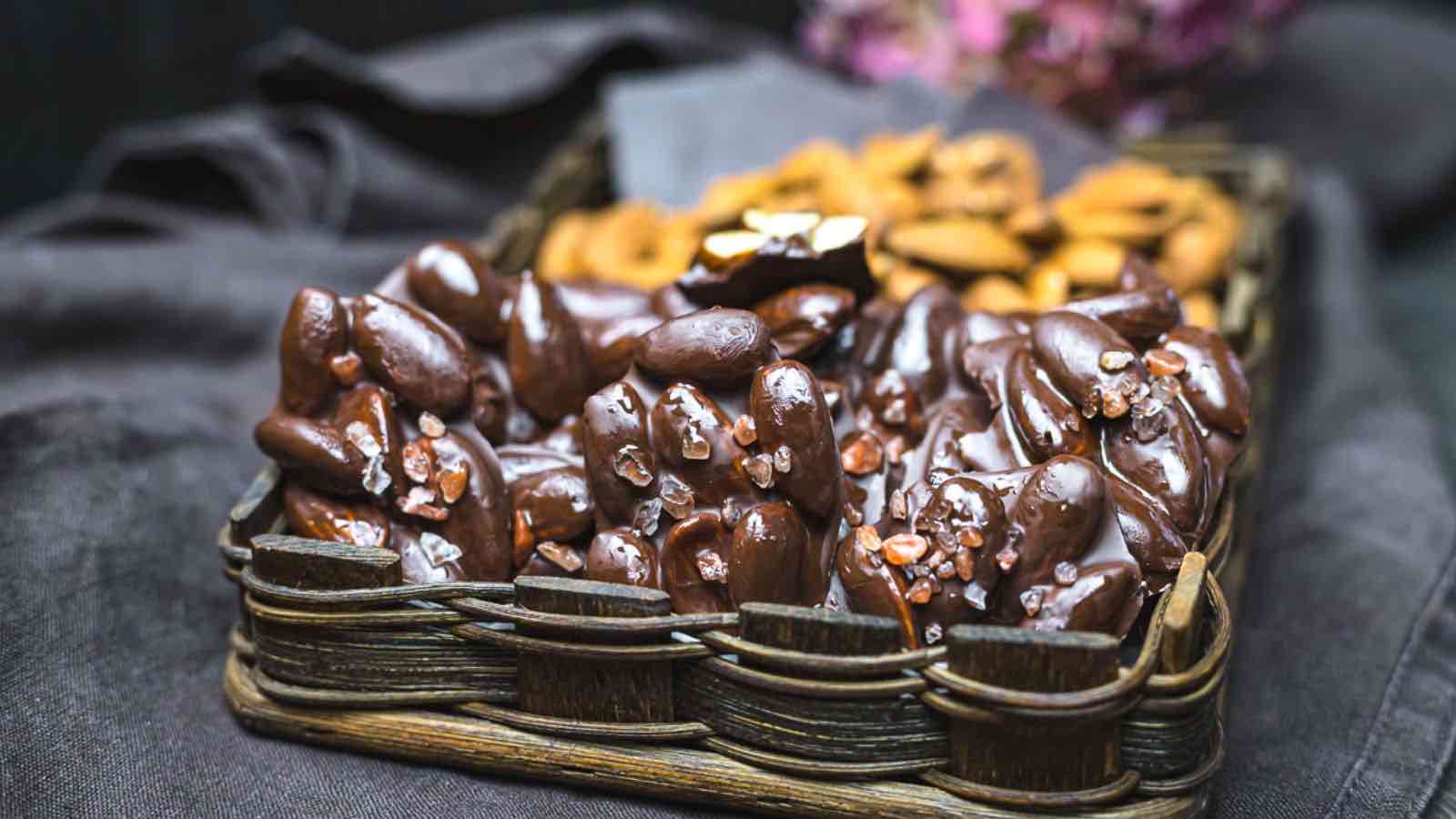 A wicker basket filled with chocolate-covered almonds sprinkled with sea salt, with dried fruit in the background.