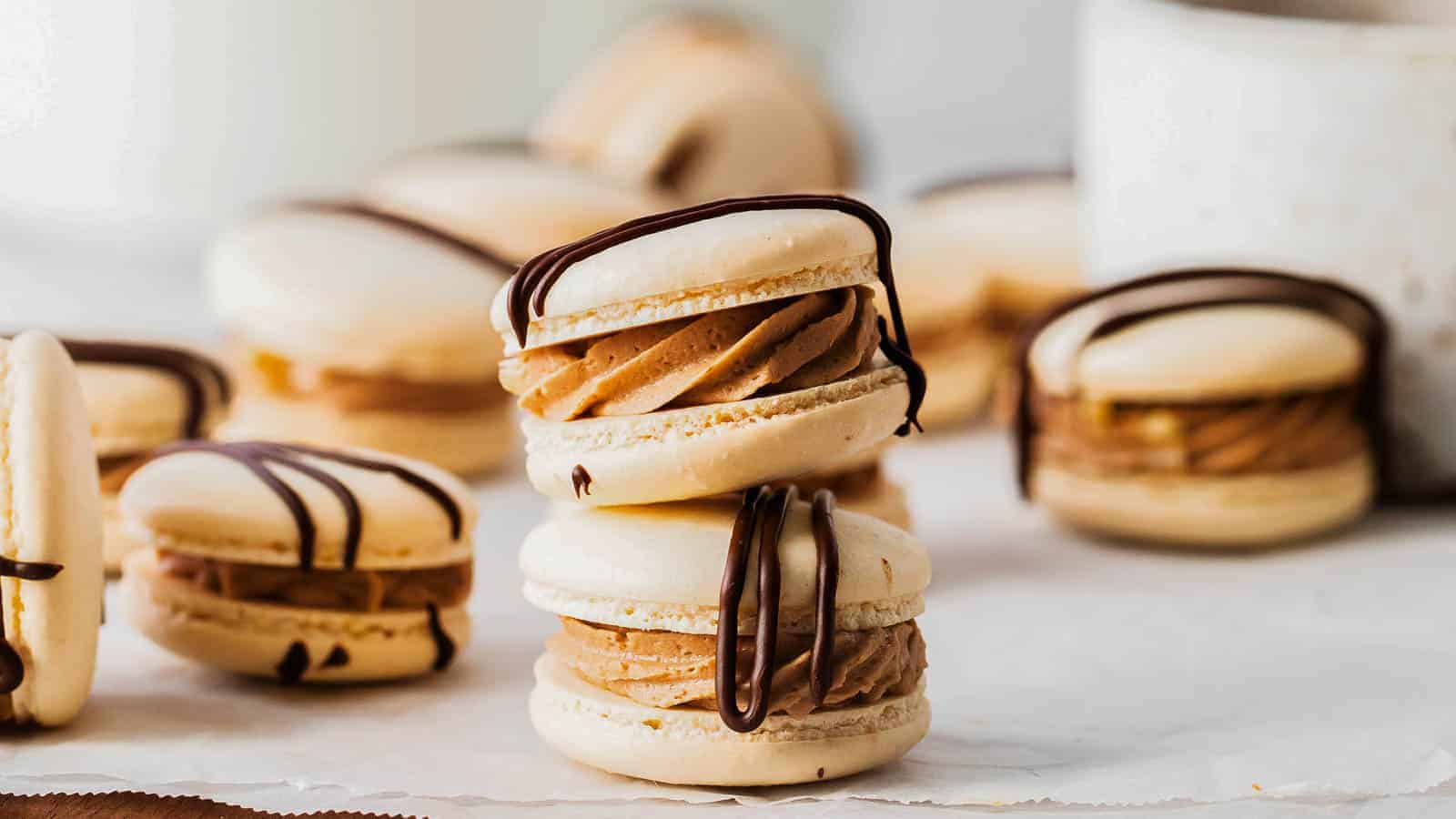 A close-up of vanilla macarons filled with brown cream and drizzled with chocolate, arranged on parchment paper with more macarons in the background.