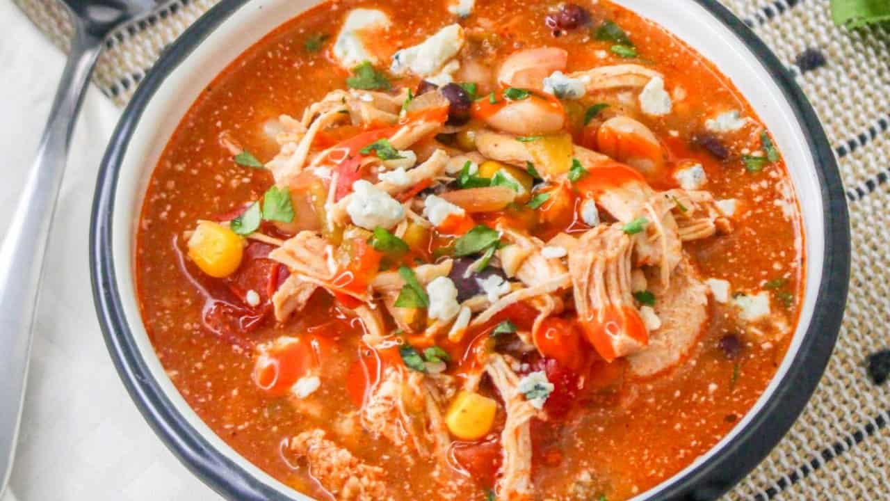 A bowl of chicken tortilla soup with shredded chicken, corn, beans, tomatoes, cheese crumbles, and fresh herbs on top.