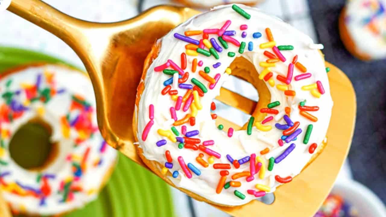 A donut with white frosting and colorful sprinkles is held by a golden spatula above a green plate.