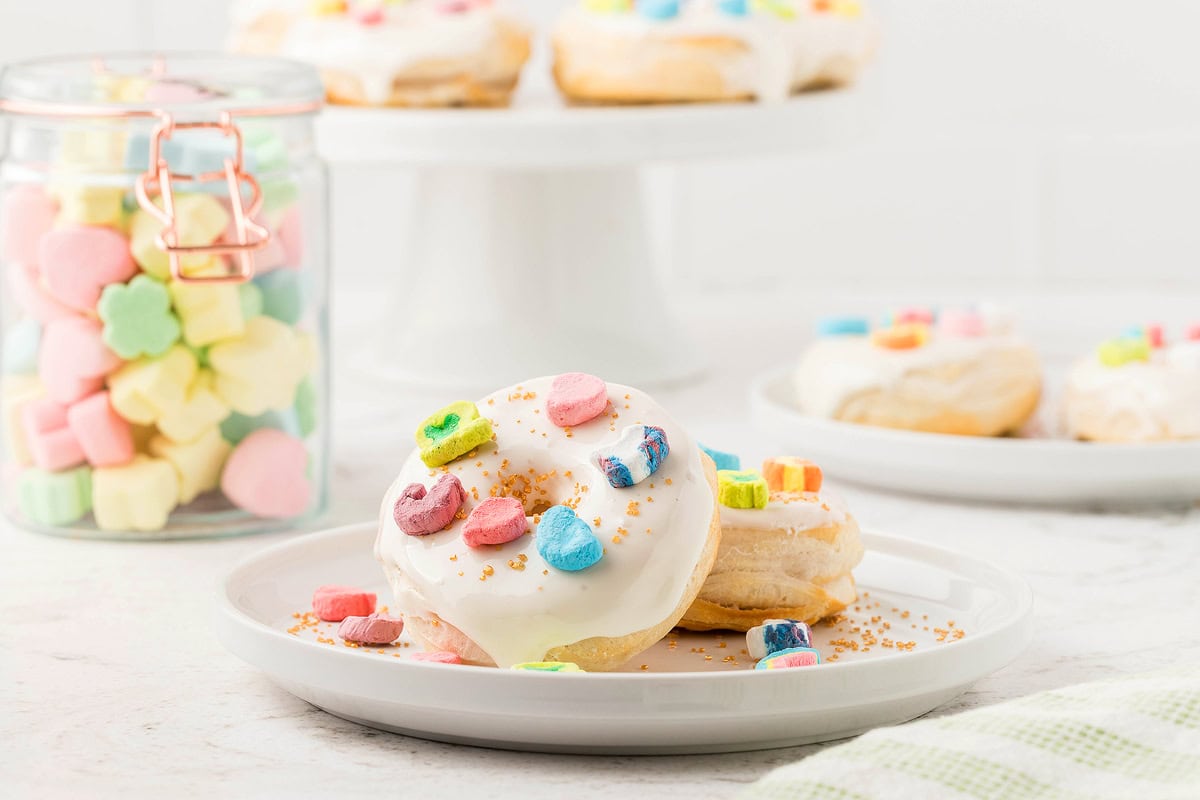 A white plate with a donut covered in white icing and colorful cereal pieces, next to a jar of pastel marshmallows and more donuts on a cake stand in the background.