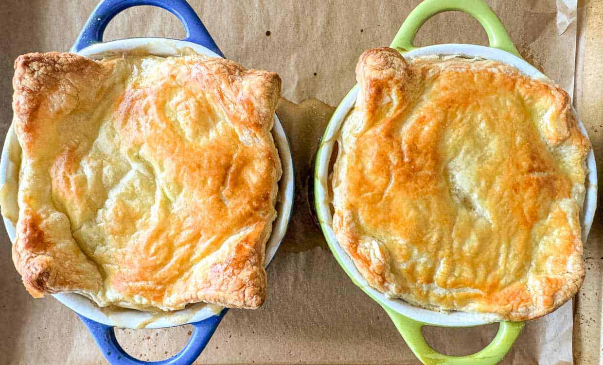 Two individual pot pies in blue and green ramekins with golden, flaky puff pastry tops on a parchment-lined baking sheet.