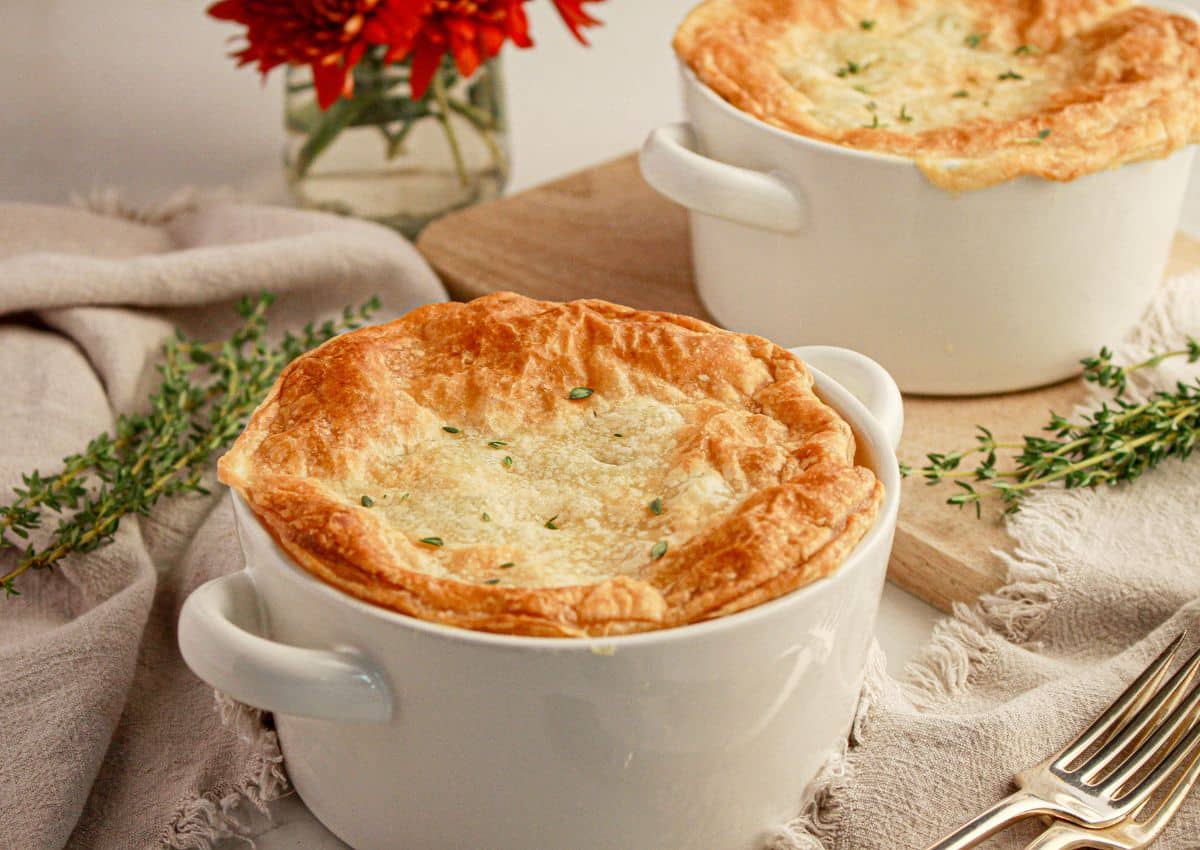 Two white ramekins filled with golden brown pot pie sit on a table, garnished with fresh thyme, next to a beige napkin, silverware, and a vase with red flowers.