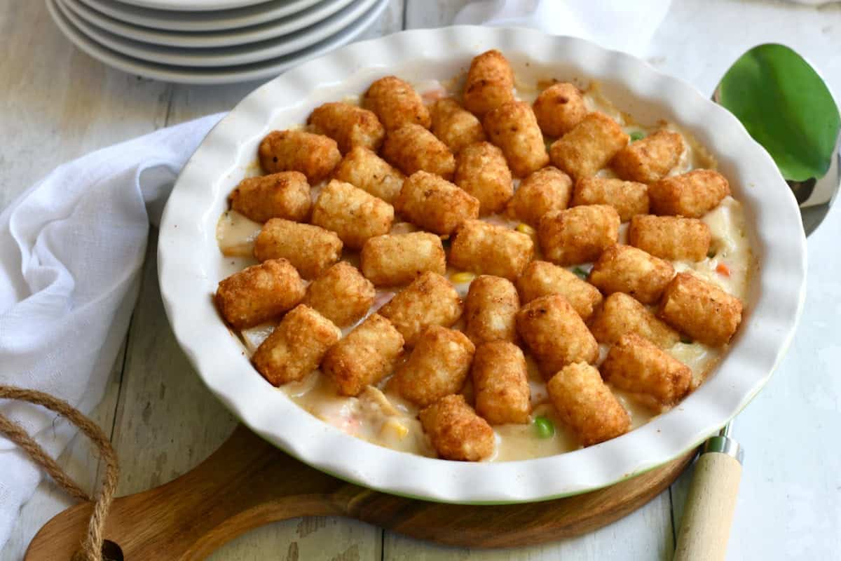 A round dish filled with a creamy casserole topped with evenly spaced tater tots, set on a wooden board with a serving spoon and a stack of plates nearby.