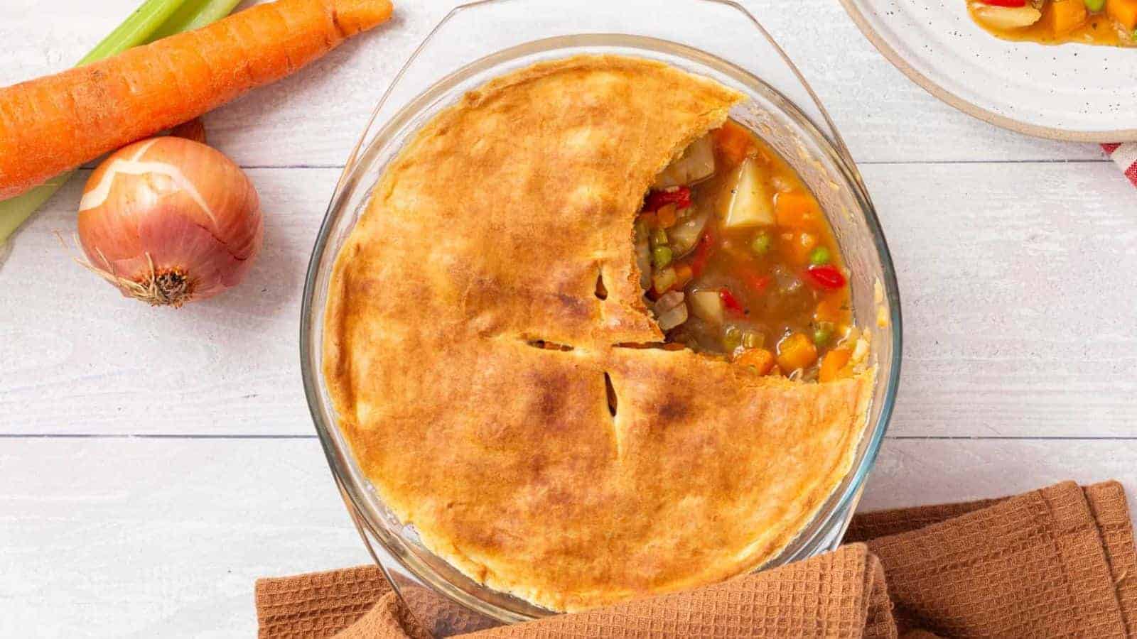 A round vegetable pot pie with a golden crust sits in a glass baking dish; a slice is missing. An onion, carrot, and napkin are nearby on a white surface.