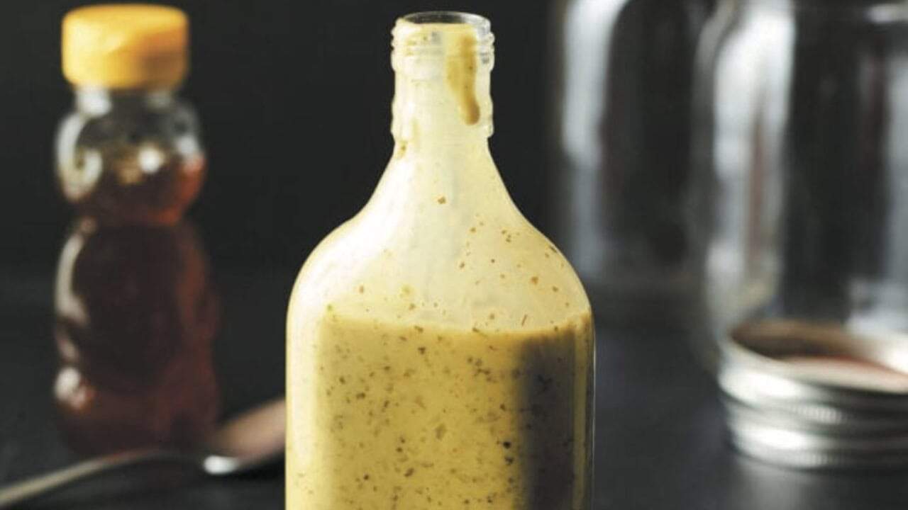 A glass bottle filled with yellow salad dressing sits on a dark surface, with a honey bear bottle and a spoon blurred in the background.