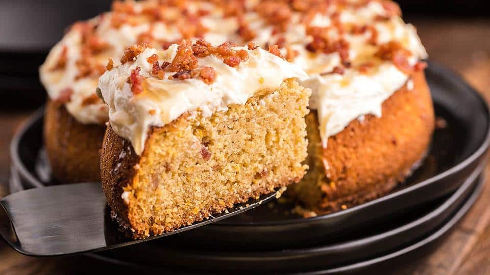 A slice of cake with white frosting and bacon bits is being lifted from a whole cake on a black plate.