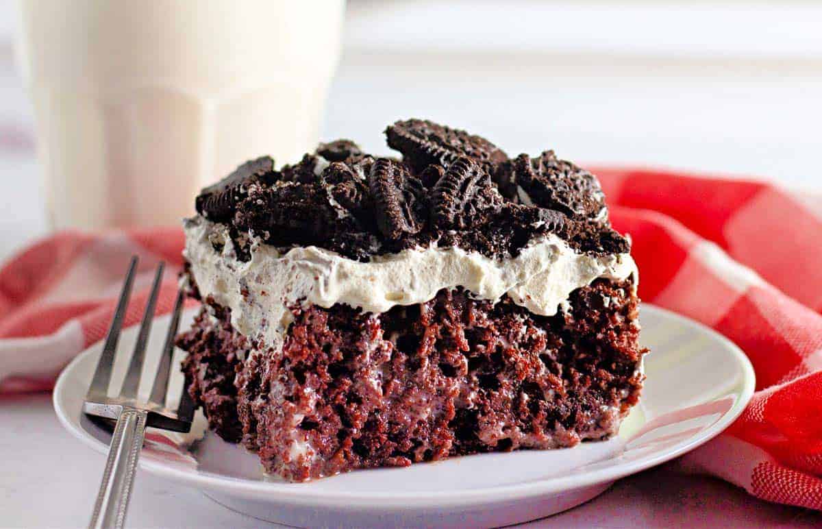 A slice of chocolate poke cake topped with whipped cream and crushed cookies on a white plate with a fork, next to a red and white napkin.