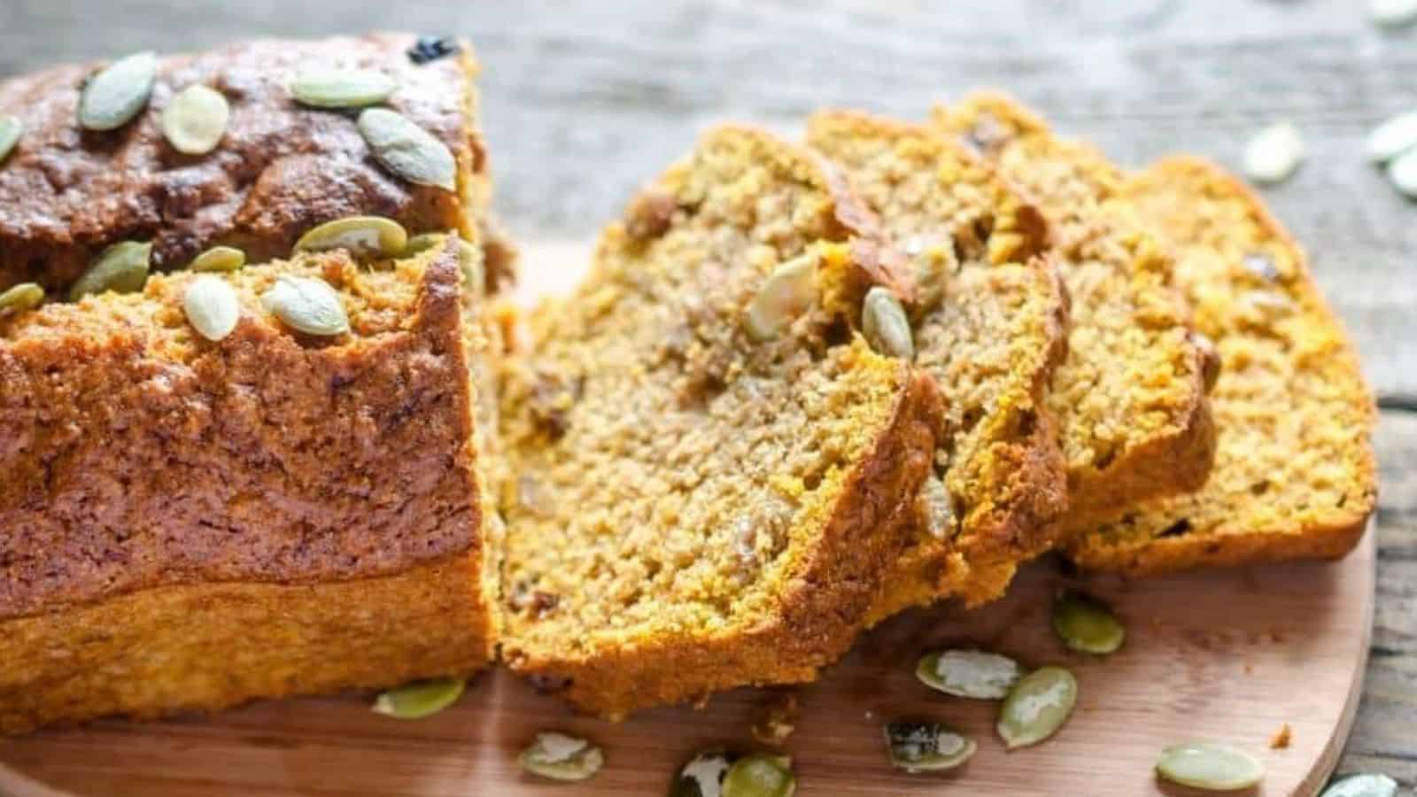 A loaf of pumpkin bread with pumpkin seeds on top, partially sliced, sits on a wooden board.