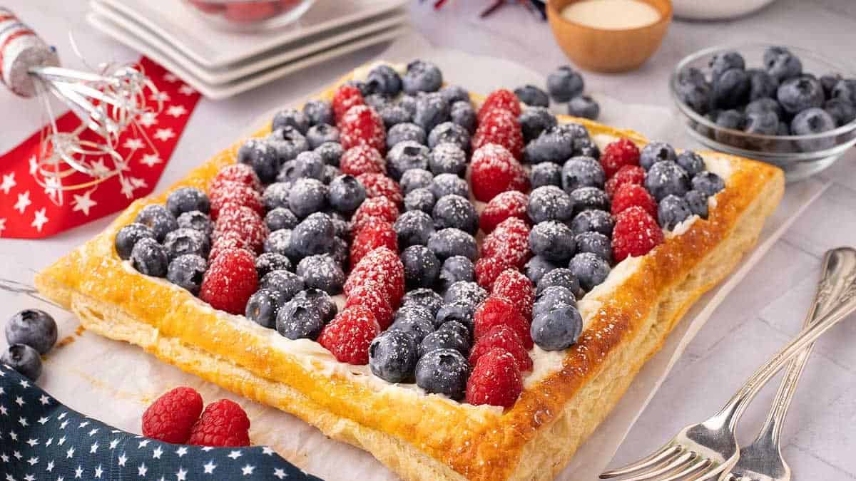 A rectangular pastry topped with rows of blueberries and raspberries, dusted with powdered sugar, sits on parchment paper with utensils and bowls nearby.