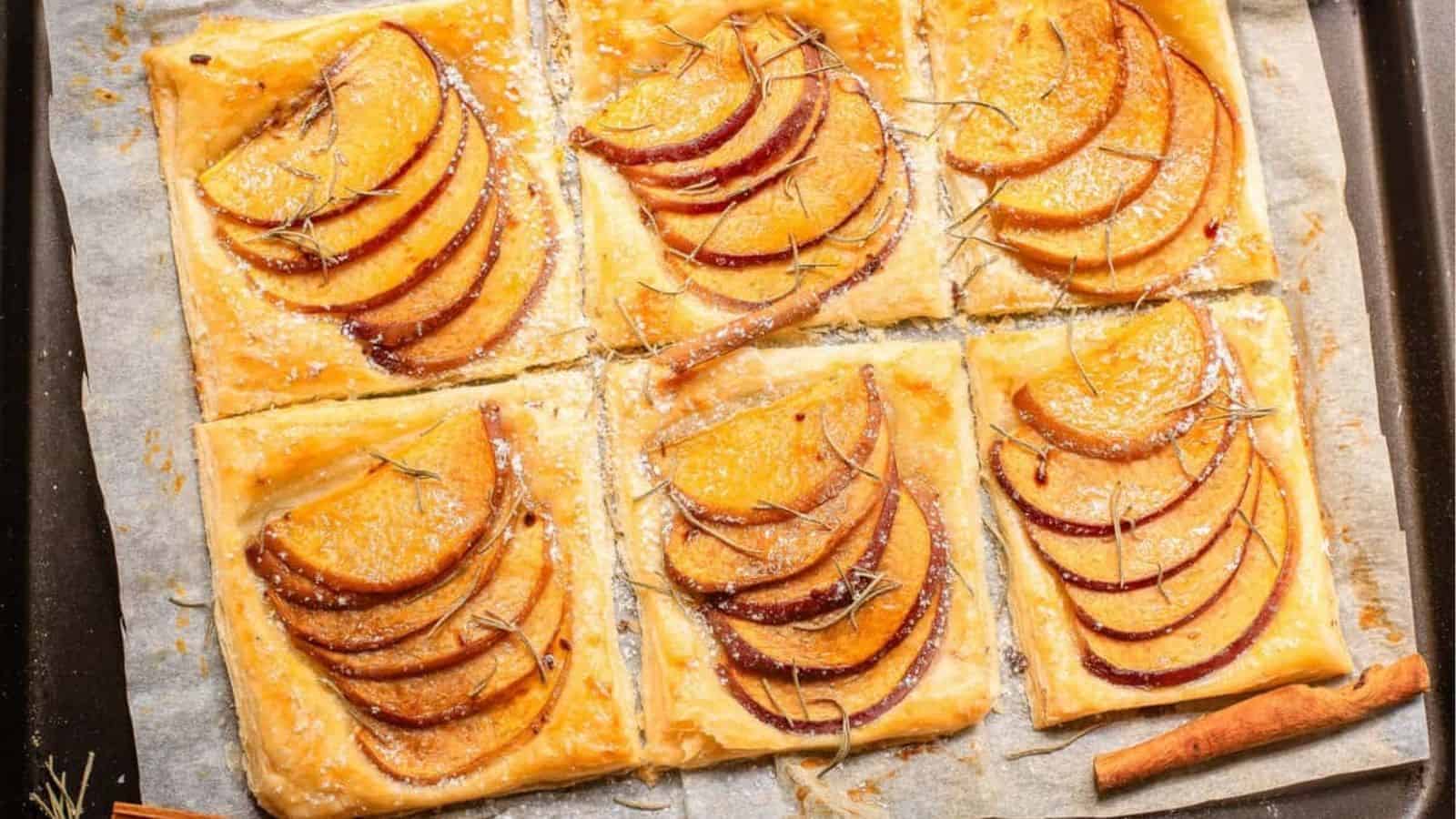 Six rectangular peach tarts with puff pastry, arranged on parchment paper, topped with peach slices and a sprinkle of herbs, next to a cinnamon stick.