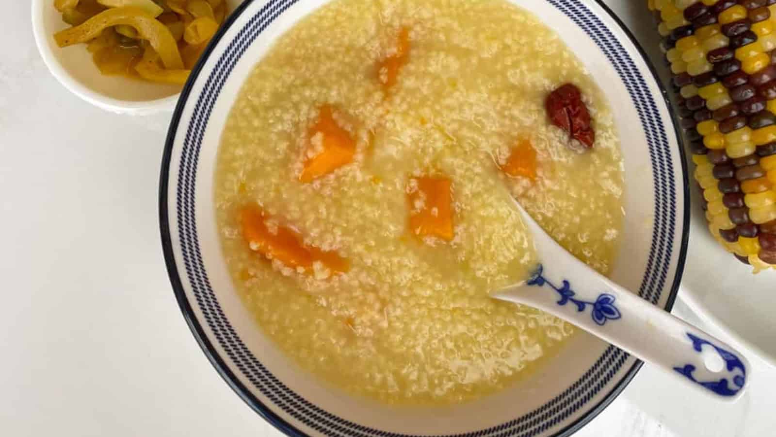 A bowl of millet porridge with orange chunks, accompanied by a spoon, sliced corn on the cob, and a small dish of pickled vegetables.