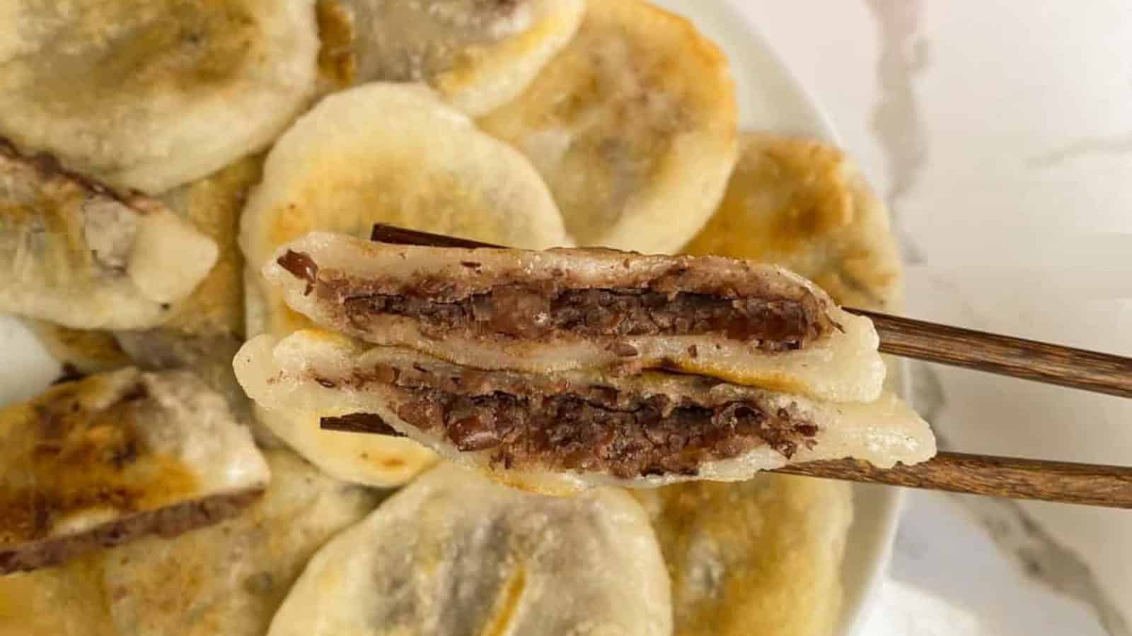 A pair of chopsticks holds a halved dumpling filled with sweet red bean paste above a plate of similar dumplings.