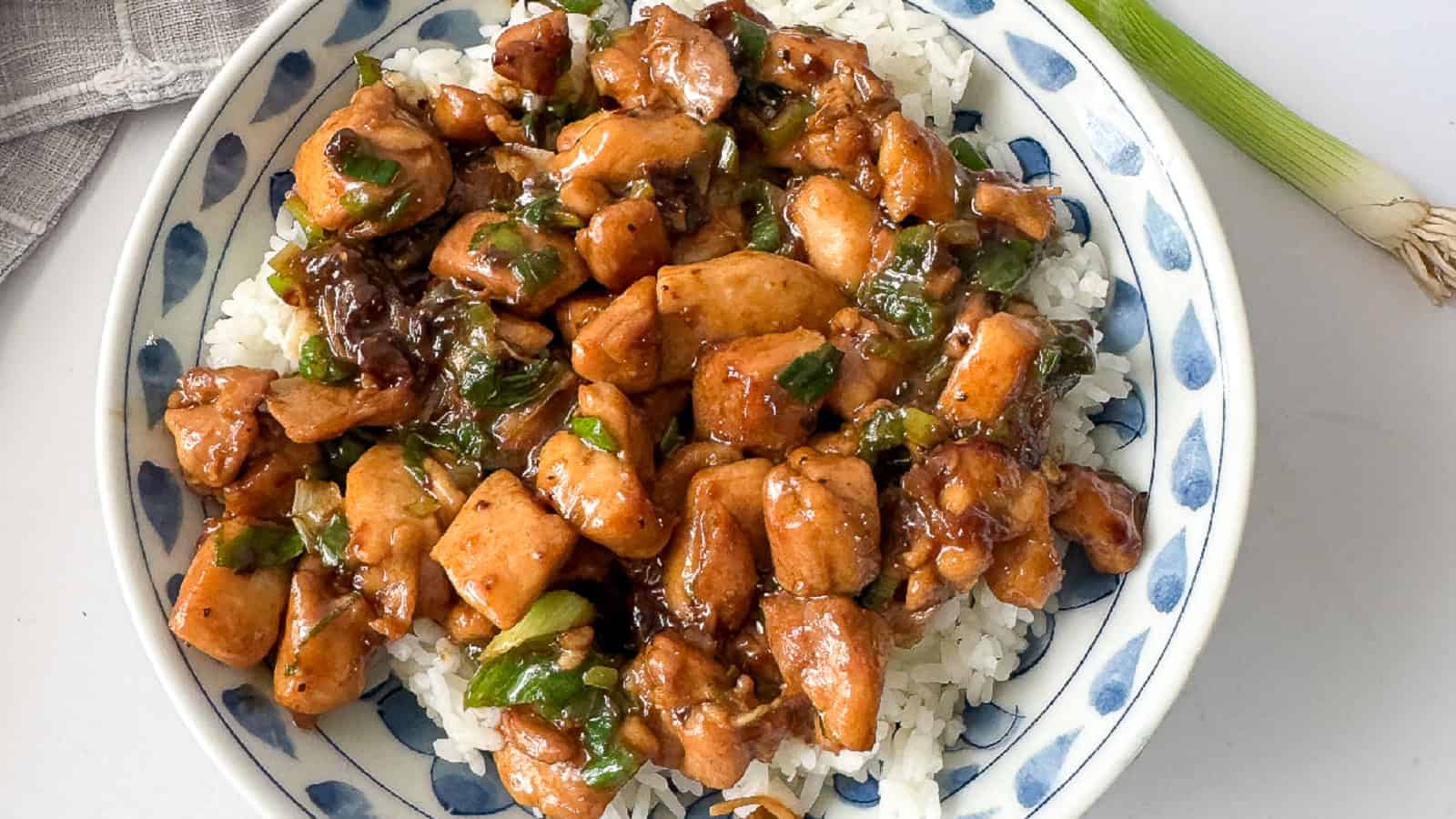 A plate of white rice topped with diced chicken cooked in a brown sauce with green onions.