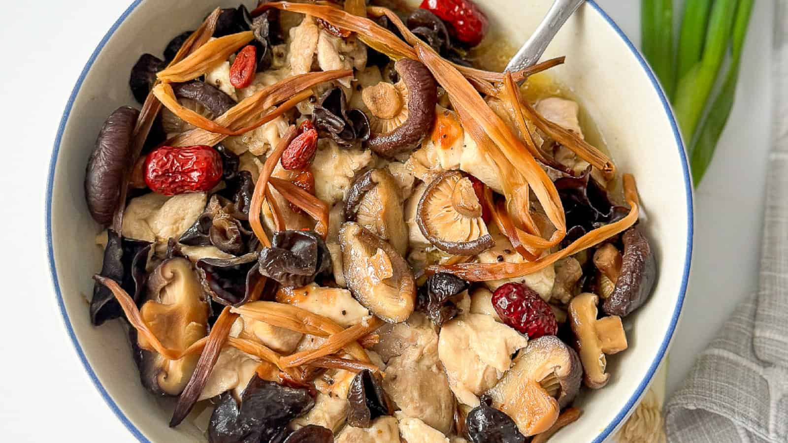 A bowl of braised chicken with mushrooms, goji berries, and dried red dates, garnished with sliced herbs.