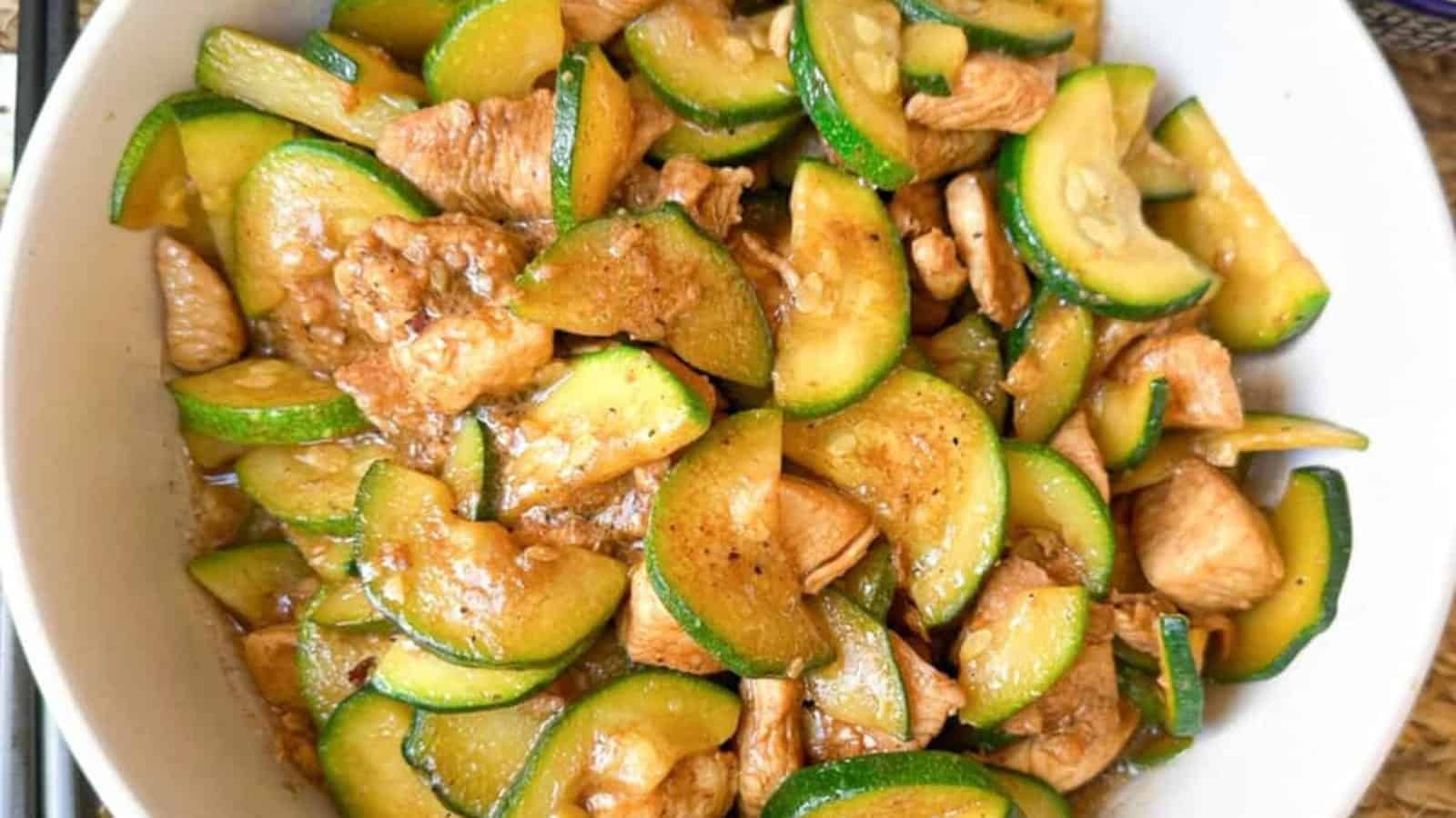 Chicken and zucchini stir fry in a white bowl, featuring sliced zucchini and pieces of cooked chicken coated in a brown sauce.