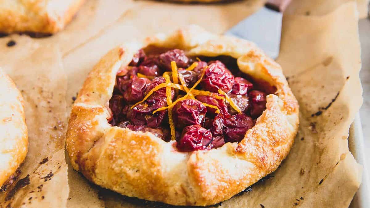 A rustic cherry galette with a golden, flaky crust, filled with cherries and topped with thin orange zest strips, sits on brown parchment paper.