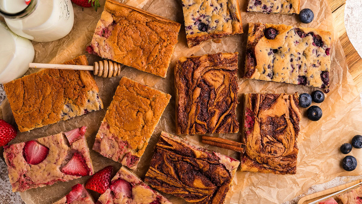 An assortment of sliced fruit and cinnamon swirl bars arranged on parchment paper, with strawberries, blueberries, honey dipper, and bottles of milk nearby.