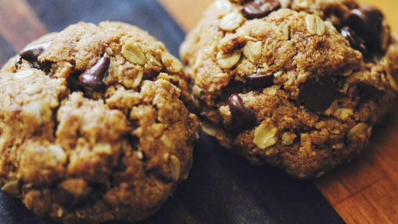 Two oatmeal chocolate chip cookies rest on a wooden surface, showing visible oats and chocolate chips in their slightly rough texture.