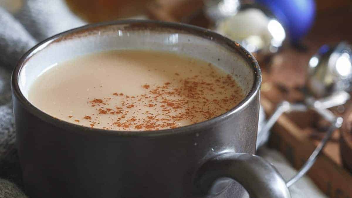 A grey ceramic mug filled with a creamy hot beverage, lightly sprinkled with cinnamon on top, sits on a surface with blurred objects in the background.