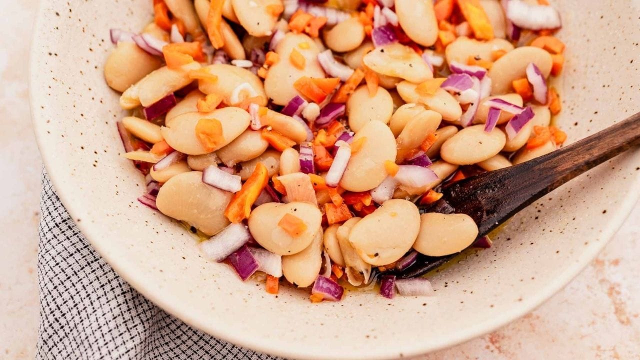 A bowl of salad with large white beans, chopped red onions, and diced carrots, mixed with a wooden spoon.