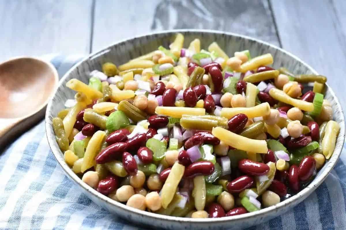 A bowl of three bean salad with kidney beans, chickpeas, yellow wax beans, celery, and red onion on a blue checkered cloth.