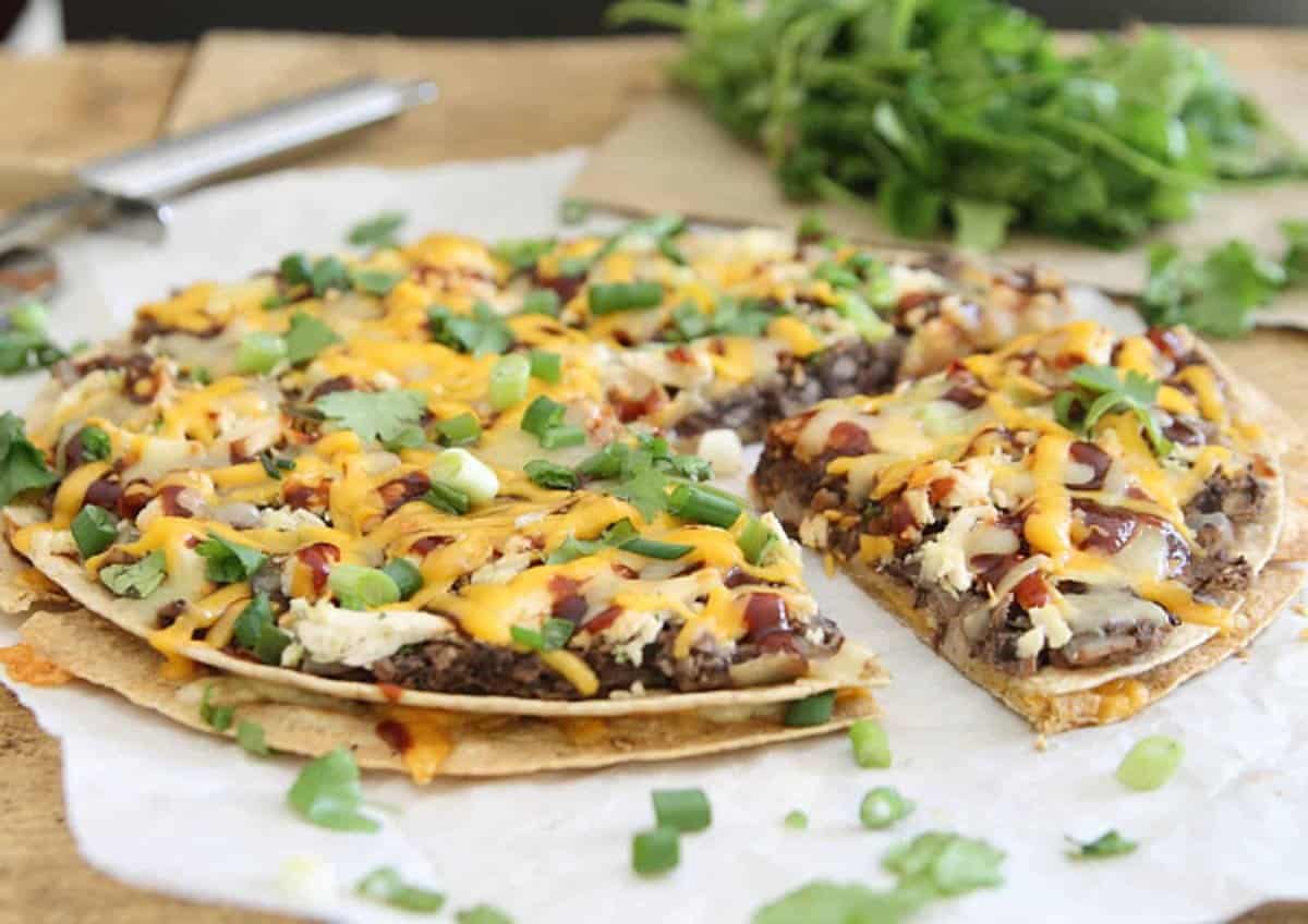 A baked flatbread pizza topped with melted cheese, green onions, and herbs sits on parchment paper with a slice missing; fresh greens are in the background.