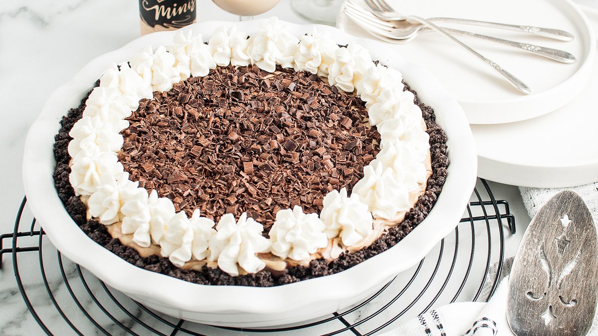 A chocolate cream pie topped with whipped cream and chocolate shavings sits on a cooling rack, with plates, forks, and a pie server nearby.