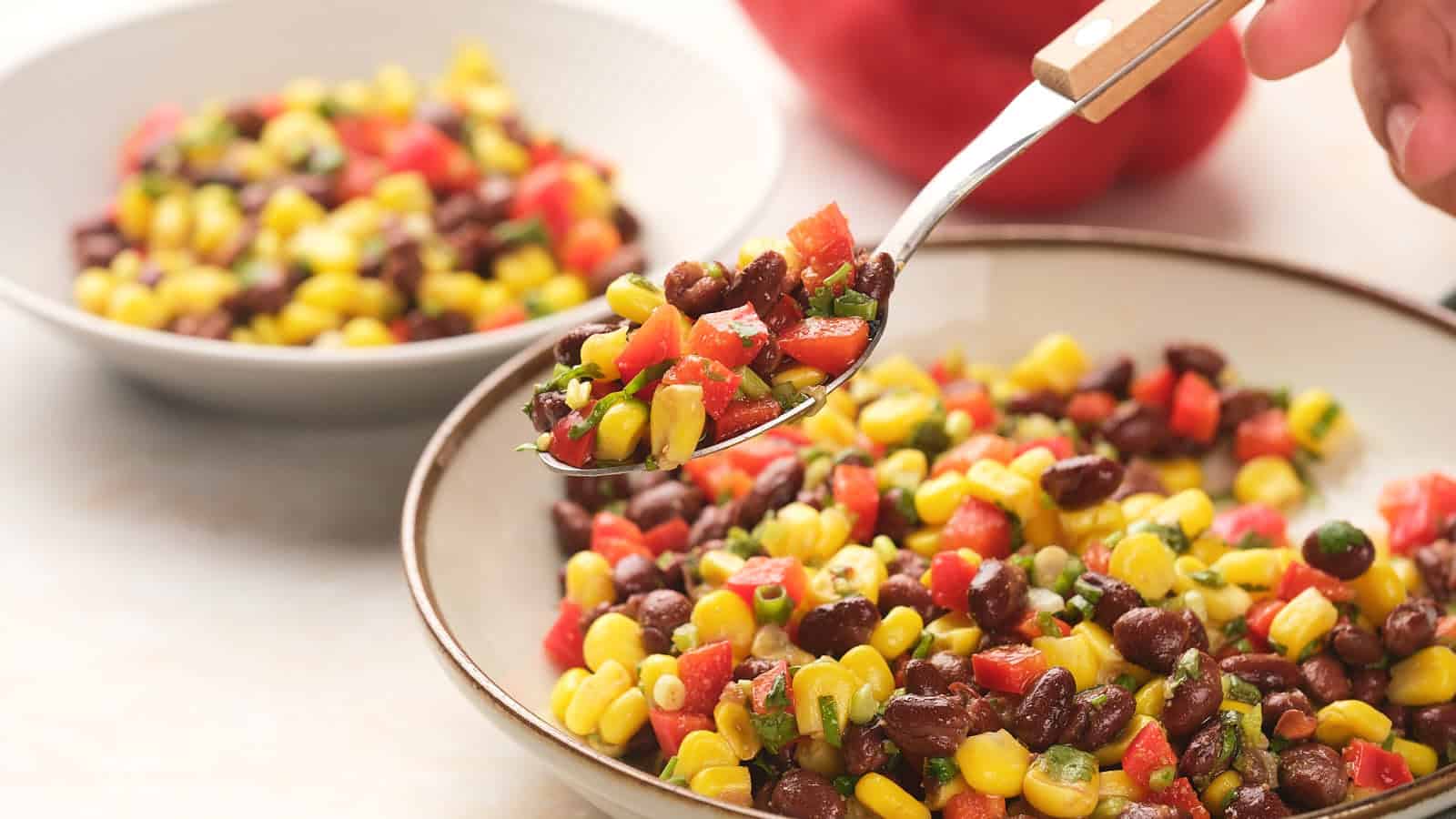 A person holds a spoonful of corn, black bean, and diced vegetable salad over a bowl, with another bowl of the same salad in the background.