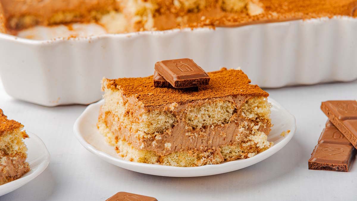A slice of chocolate tiramisu topped with chocolate pieces on a white plate, with a baking dish and chocolate squares in the background.