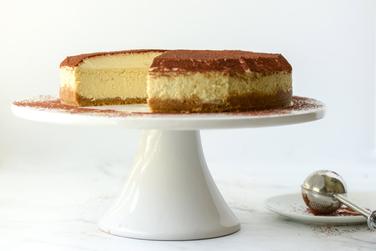 A cheesecake with a slice removed sits on a white cake stand, with a metal ice cream scoop and a plate in the background.