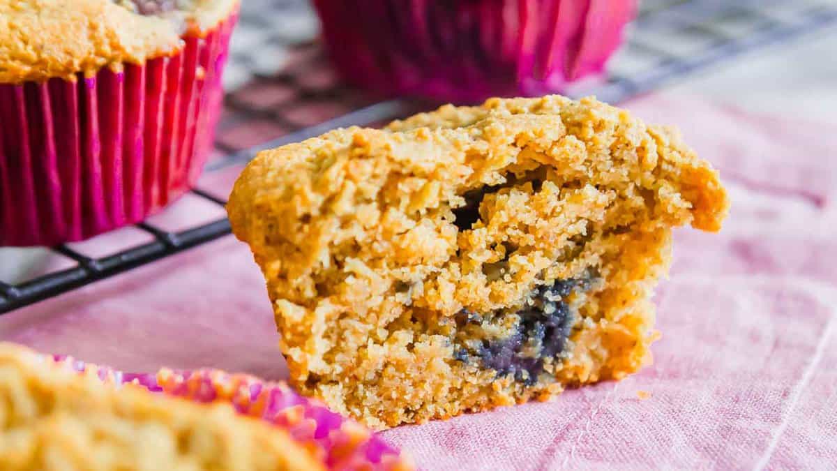 A close-up of a halved blueberry muffin with a crumbly texture, sitting on a pink cloth, with more muffins on a cooling rack in the background.
