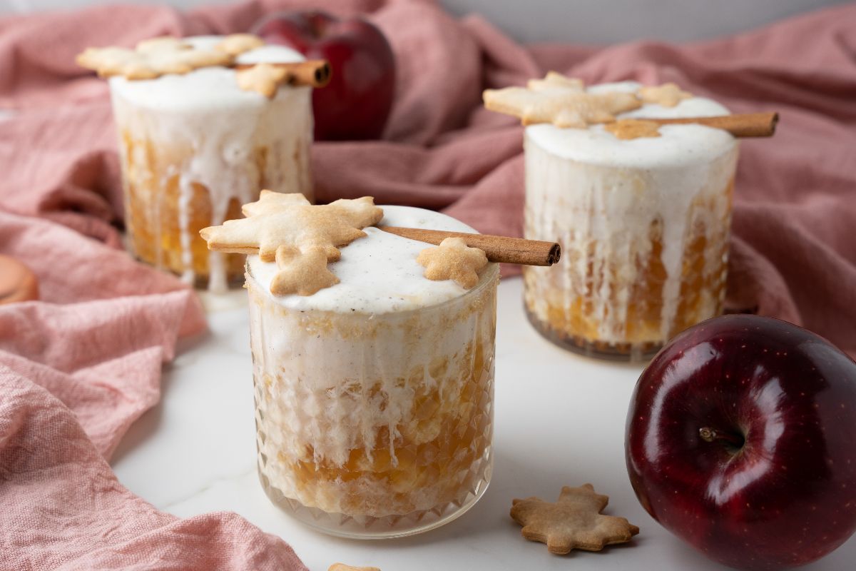Three glasses filled with layered dessert topped with foam, cinnamon sticks, and leaf-shaped cookies; red apples and a pink cloth in the background.
