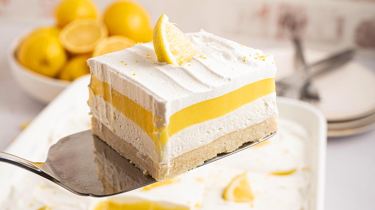 A dessert bar with a crumbly base, a yellow lemon layer, and a thick white whipped topping, garnished with a lemon wedge on top of a serving spatula.