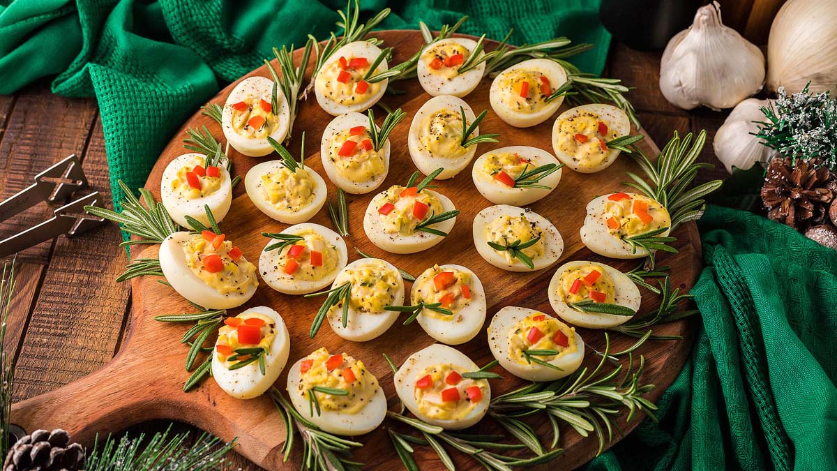 A wooden serving board with deviled eggs topped with diced red and yellow peppers and rosemary sprigs, surrounded by more rosemary, with green linens and garlic in the background.