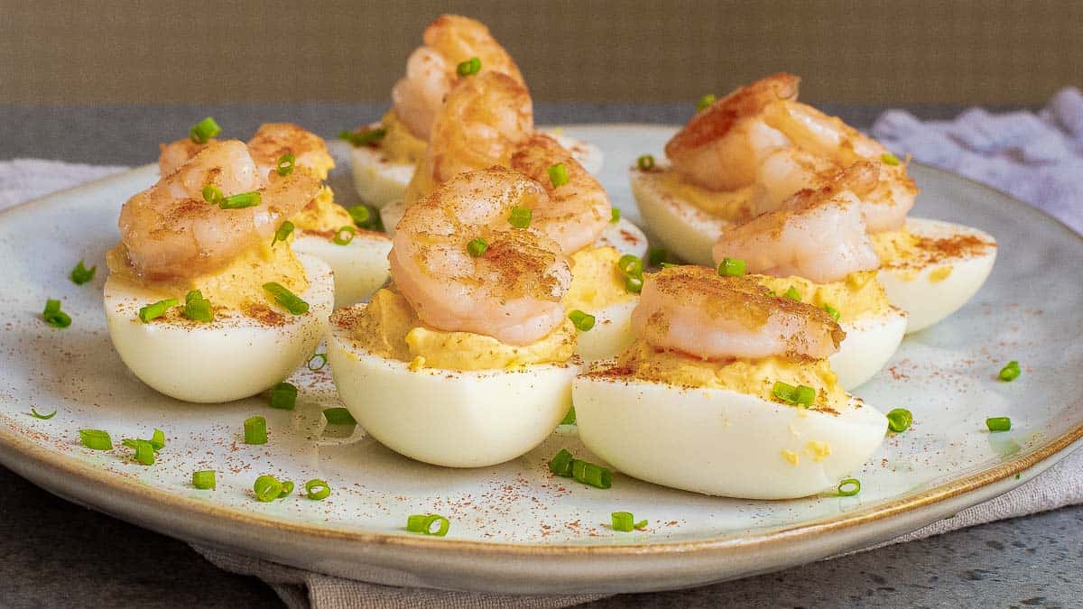 A plate of deviled eggs topped with cooked shrimp and sprinkled with chopped chives and paprika.
