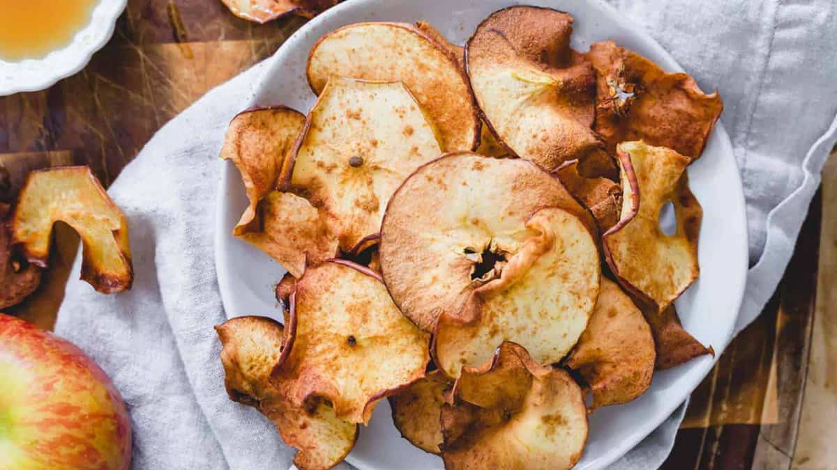 A white plate filled with baked apple chips sits on a white cloth, with a fresh apple and more chips visible nearby.