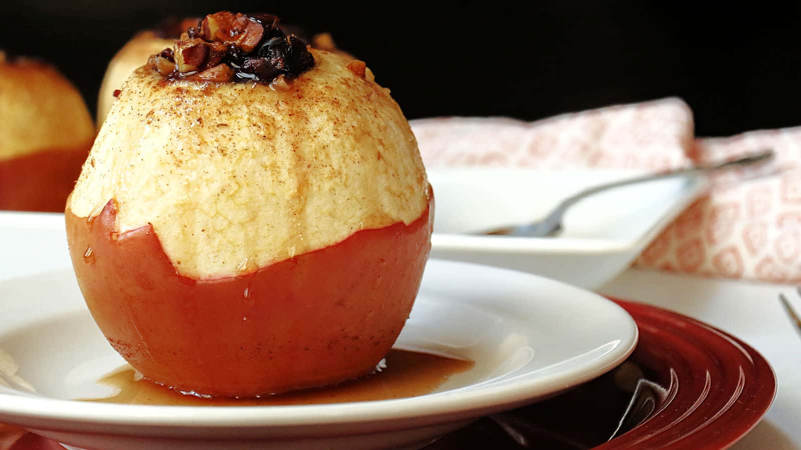 A baked apple with a peeled top sits on a white plate, filled with chopped nuts and spices, with syrup pooling around the base.