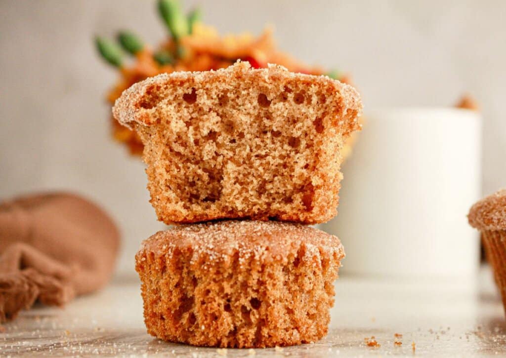 Two cinnamon sugar muffins are stacked, with the top muffin cut in half to show a fluffy, moist interior. A blurred mug and fall decor are in the background.