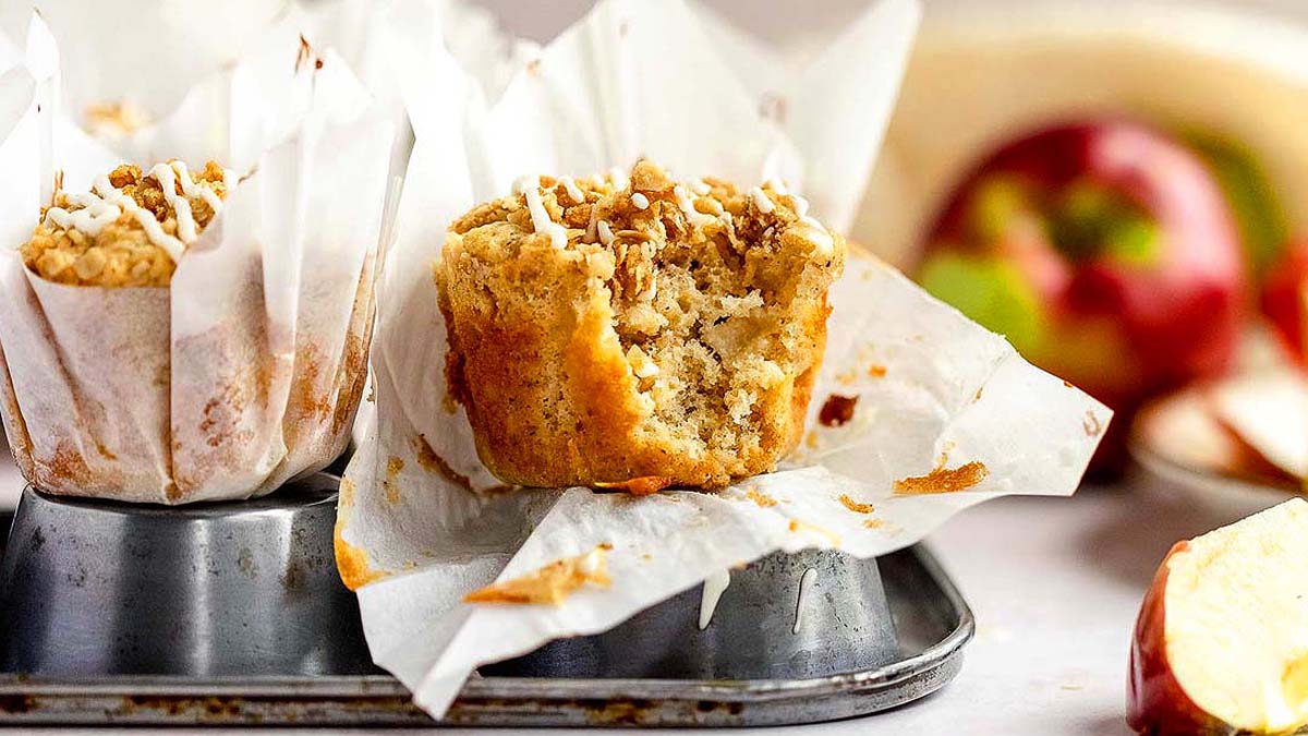 A partially eaten muffin with a crumb topping sits in a parchment liner on a muffin tin, with apples and more muffins in the background.