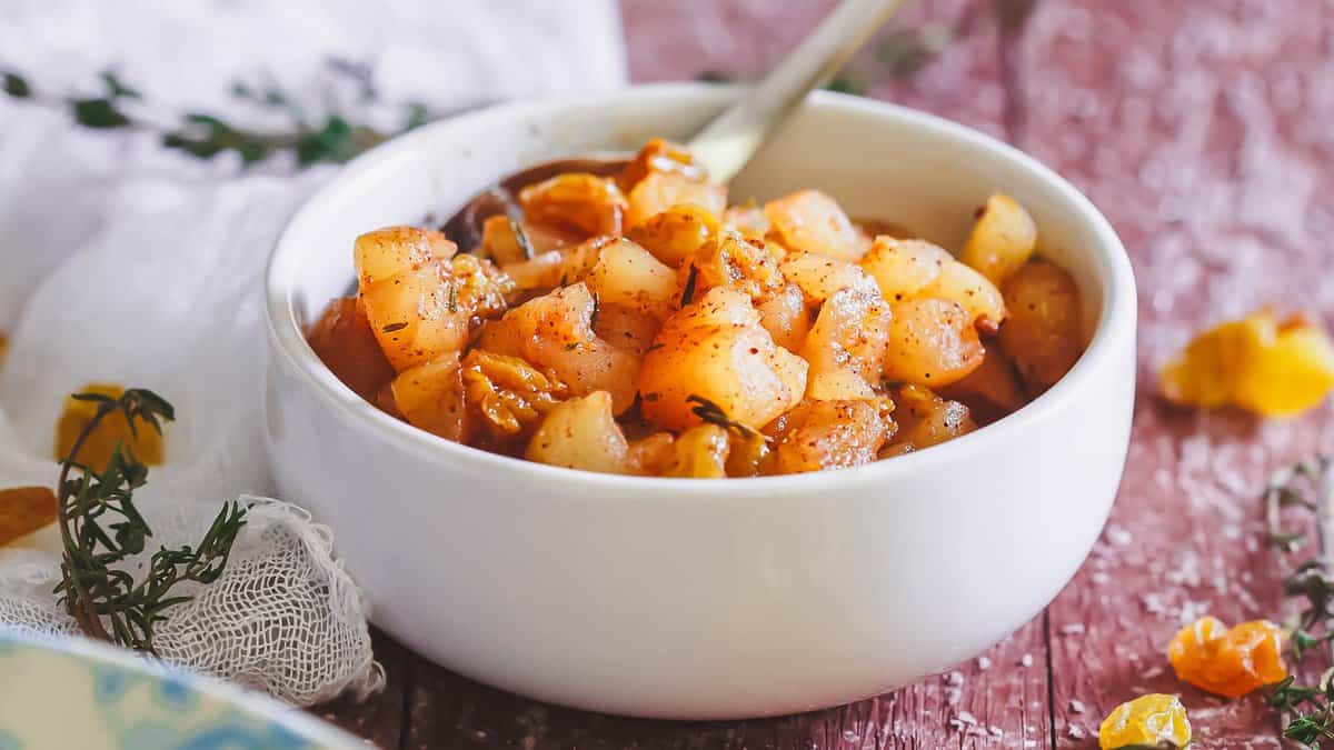 A white bowl filled with cooked, diced apples seasoned with spices, with a spoon inside the bowl, placed on a rustic wooden surface.