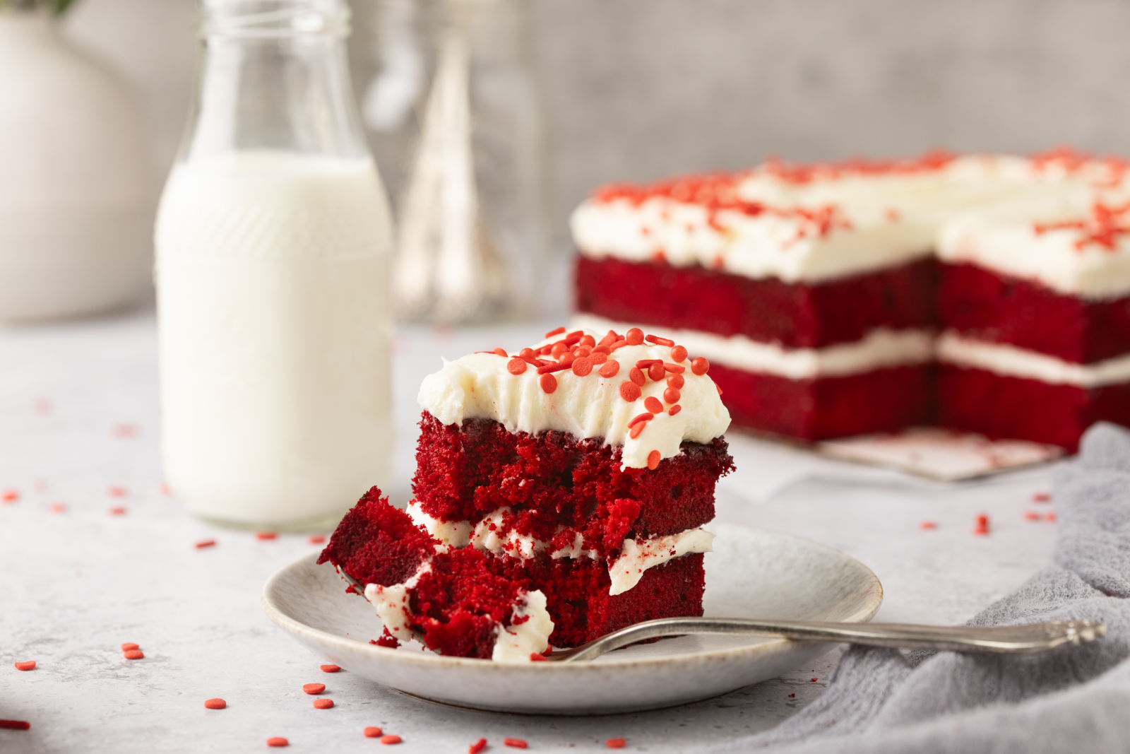 A slice of red velvet cake with cream cheese frosting and red sprinkles on a plate with a fork, a bottle of milk, and the remaining cake in the background.