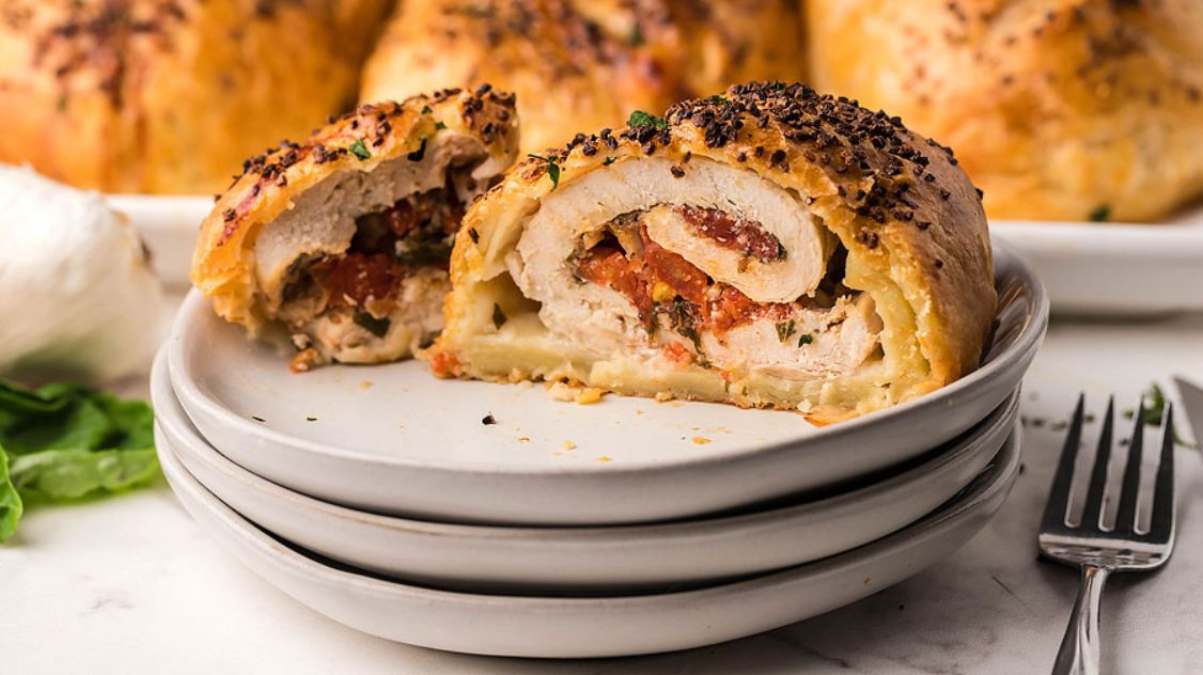 Sliced stuffed chicken breast wrapped in golden pastry on stacked plates, revealing a filling with tomatoes and herbs. A fork is beside the plates.