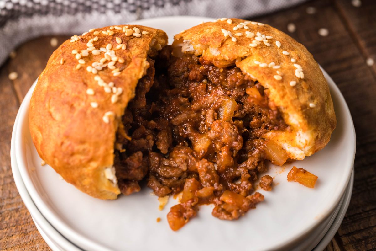 A golden brown meat-filled bun with sesame seeds, split open to reveal a savory ground meat and vegetable filling, served on a white plate.