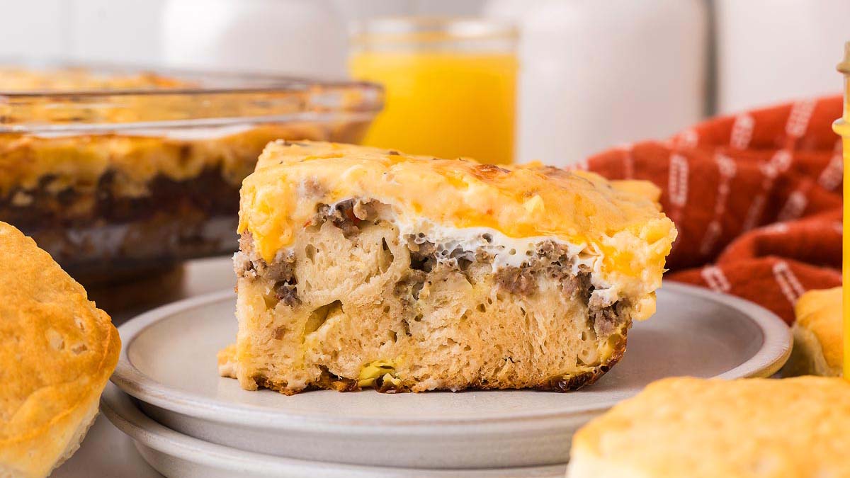 A slice of breakfast casserole with sausage, eggs, and cheese on a plate, surrounded by biscuits, with a glass of orange juice in the background.