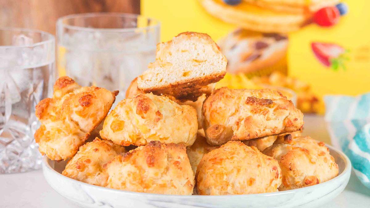 A plate of golden-brown cheese biscuits is stacked next to two glasses of ice water, with a yellow box and a striped napkin in the background.