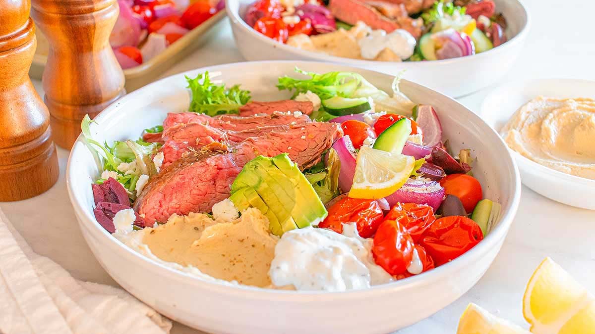 A bowl with sliced steak, avocado, tomatoes, cucumber, onions, mixed greens, feta cheese, hummus, and tzatziki sauce. Another similar bowl and condiments are in the background.