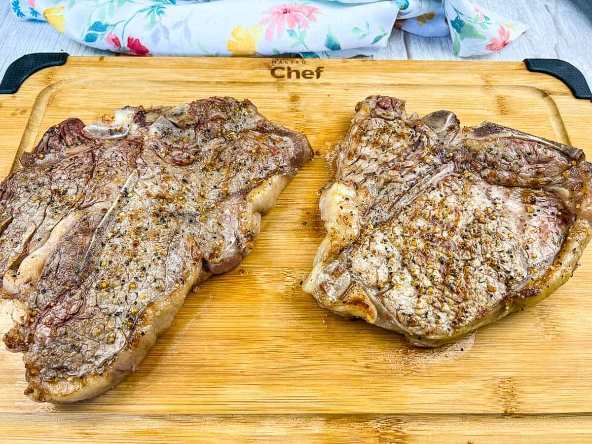 Two cooked T-bone steaks seasoned with pepper and spices sit on a wooden cutting board with a floral cloth in the background.