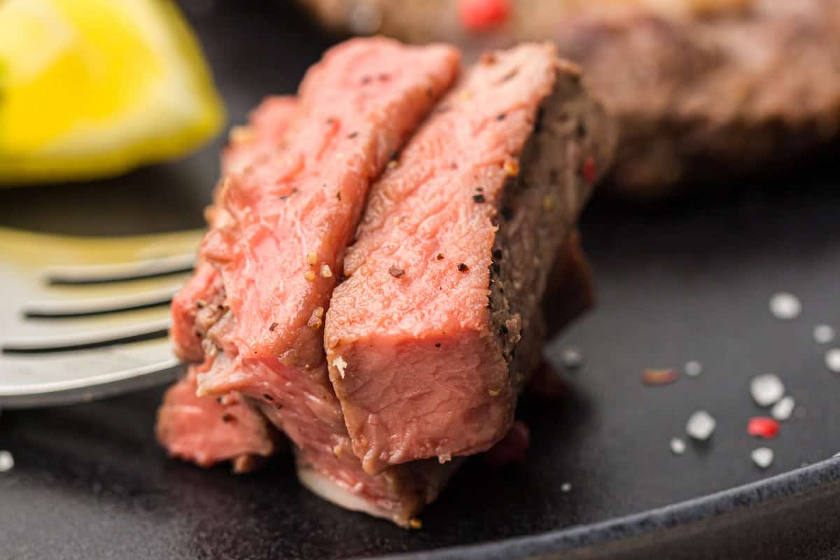 Two slices of medium-rare steak, seasoned with pepper, are shown on a black plate with a metal fork and a lemon wedge in the background.